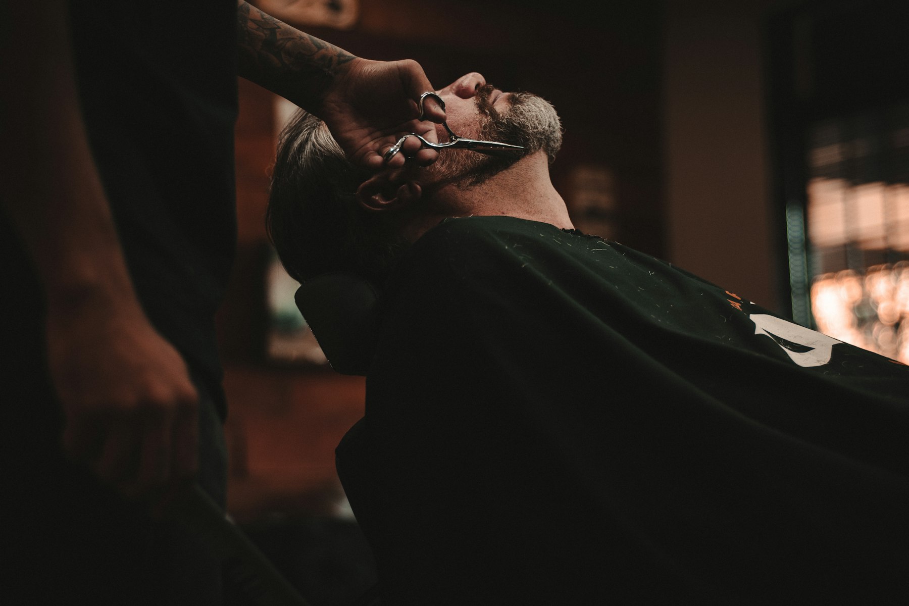 A barber trimming a client's beard in a dark, lamp-lit barbershop
